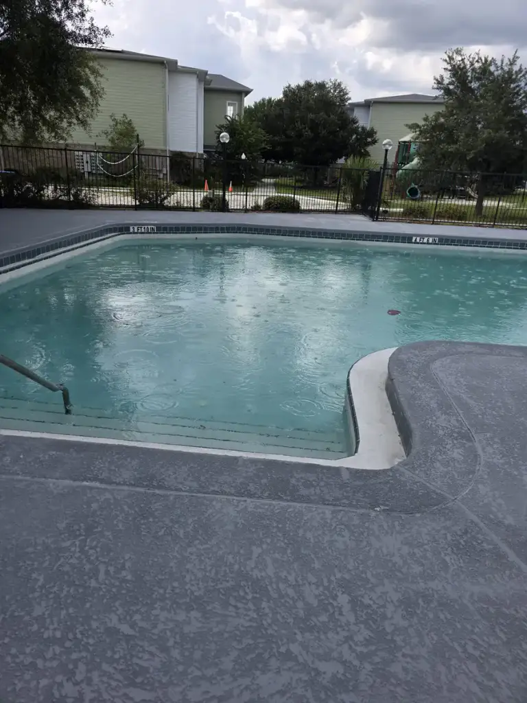 Wide completed view of the gray pool deck, blue water, fence, and community building.